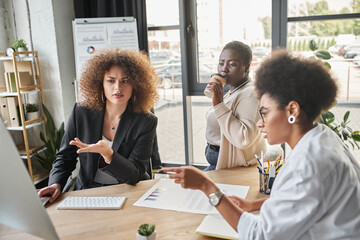 diverse group of multiethnic businesswomen talking near computer and documents with graphs in office
