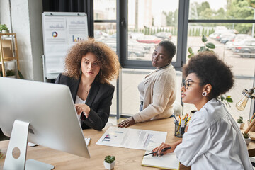 diverse group of multiethnic businesswomen talking near computer and documents with graphs in office