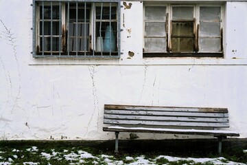 Alte braune verwitterte Holzbank mit Schnee auf Wiese vor alter weißer rissiger Hausfassade mit zwei Fenstern und Eisengittern am Nachmittag im Winter