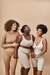 smiling plus size multiracial women in lingerie posing on beige, beauty and body positivity