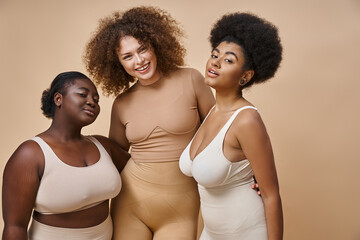 happy multiethnic plus size girlfriends in lingerie posing on beige backdrop, body positive beauty