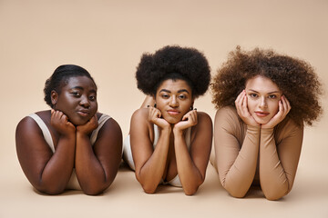 charming multiethnic body positive women in lingerie lying down and looking at camera on beige