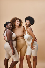 happy multiethnic body positive women in underwear embracing on beige, natural curvy beauty