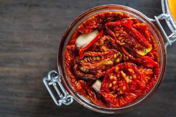 Sun-dried red tomatoes with garlic, green rosemary, olive oil and spices in a glass jar on a wooden table. Rustic style, top view, closeup