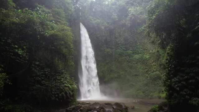 Large waterfall in deep green canyon, camera fly low and slowly towards power stream of water rushing down. Tropical plants hang from vertical walls of gorge, water mist in the air