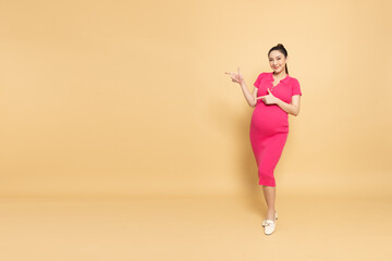 Happy Asian Pregnant woman standing smile and pointing to copy space isolated on brown background, Motherhood and Pregnancy Concept, Full body compostion