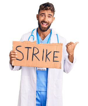 Young hispanic man wearing doctor stethoscope holding strike banner pointing thumb up to the side smiling happy with open mouth