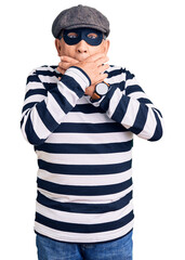 Senior handsome man wearing burglar mask and t-shirt shocked covering mouth with hands for mistake. secret concept.