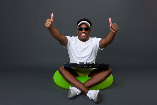 An African man with a smile on his face wearing dark glasses sits in a green inflatable circle with a Dij console, giving thumbs up. Satisfied and happy DJ with equipment resting on a gray background