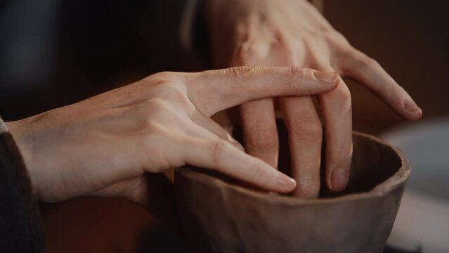 Close-up female hands making handmade cup or small vessel vase. Woman potter wearing warm plaid shirt shapes craft clay tableware in pottery studio. Working process modeling in ceramic workshop