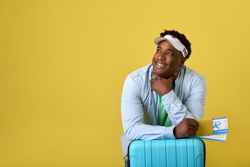 On yellow background African man in denim shirt and cap holds plane tickets in hand, looking into distance. A black man is traveler with blue suitcase waiting for announcement of boarding a flight