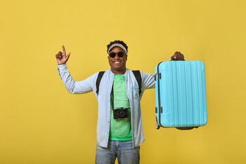Happy young African man in sunglasses holding a large blue suitcase in his hand, pointing his finger up. Dark-skinned male traveler model demonstrates a roomy suitcase for trips on a yellow background - obrazy, fototapety, plakaty