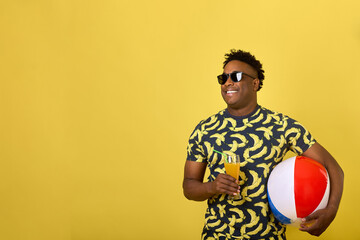 Happy dark-skinned man enjoys drinking a delicious cocktail, holding an inflatable swimming ball. On a yellow background, a young African quenches his thirst with a drink in a glass glass