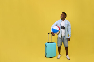 An African-American male traveler stands with a large blue suitcase and an inflatable multi-colored ball. A young modern photographer went on vacation, cheerfully looking away