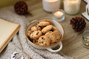 Bowl of cookies, cup of tea, dry oranges, pine cones, books, reading glasses, small presents, various Christmas decorations and lit candles on the table. Cozy Christmas hygge. Selective focus.