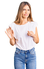 Beautiful caucasian woman with blonde hair wearing casual white tshirt disgusted expression, displeased and fearful doing disgust face because aversion reaction. with hands raised