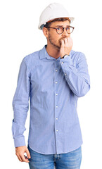 Handsome young man with bear wearing architect hardhat looking stressed and nervous with hands on mouth biting nails. anxiety problem.