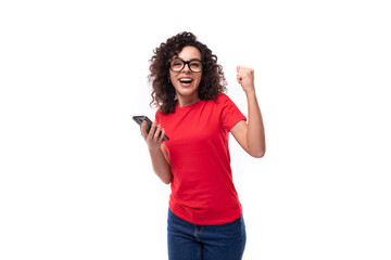 Obraz premium corporate clothing concept. young smart curly brunette female student dressed in a red t-shirt rejoices holding a smartphone in her hand