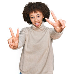 Young hispanic girl wearing wool winter sweater smiling with tongue out showing fingers of both hands doing victory sign. number two.