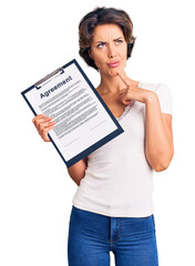 Young beautiful woman holding clipboard with agreement document serious face thinking about question with hand on chin, thoughtful about confusing idea