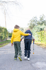 Children walking down the street, one with crutches and the other helping him