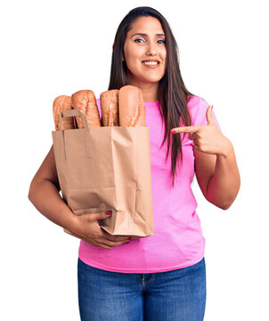 Young beautiful brunette woman holding delivery bag with bread smiling happy pointing with hand and finger