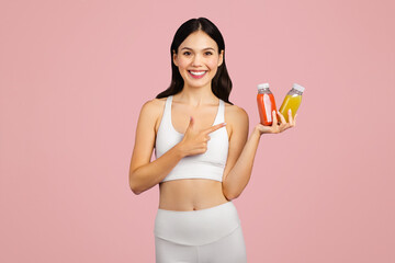 Positive caucasian lady in sportswear holding and pointing at jars of juice or smoothies isolated on pink background - obrazy, fototapety, plakaty