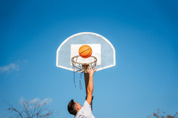 Chico joven caucásico de espaldas, con camiseta blanca saltando y ganastando una pelota de básquetball a una ganasta de metal y el cielo azul despejado en un día soleado.
