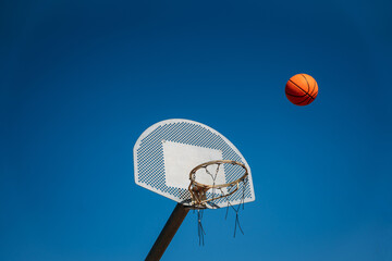Basketball basket seen from below and one side while an orange ball is falling from above. Contrasted with a blue sky on a sunny, clear day.