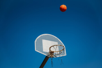 Basketball basket seen from below and one side while an orange ball is falling from above. Contrasted with a blue sky on a sunny, clear day.