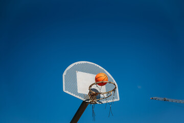 Basketball basket seen from below and one side while an orange ball is falling from above. Contrasted with a blue sky on a sunny, clear day.