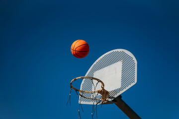 Basketball basket seen from below and one side while an orange ball is falling from above. Contrasted with a blue sky on a sunny, clear day.