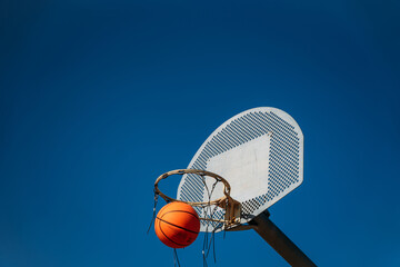 Basketball basket seen from below and one side while an orange ball is falling from above. Contrasted with a blue sky on a sunny, clear day.