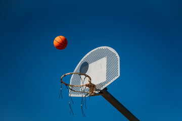 Basketball basket seen from below and one side while an orange ball is falling from above. Contrasted with a blue sky on a sunny, clear day.