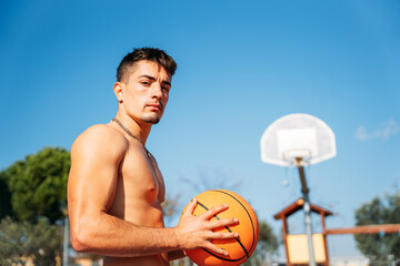 Muscular young shirtless Caucasian guy seen from close up while holding a basketball on a basketball court with net seen in the background, looking at camera.