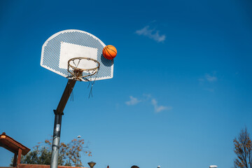 Basketball basket seen from below and one side while an orange ball is falling from above. Contrasted with a blue sky on a sunny, clear day.