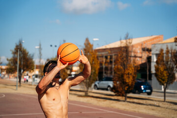 Muscular young caucasian guy without shirt seen from close up and faces while throwing a basketball on a basketball court.