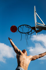 Young caucasian guy seen from below, muscular shirtless while jumping and hitting a basketball on a metal rack and clear blue sky on a sunny day. Dress the silhouette with open arms.