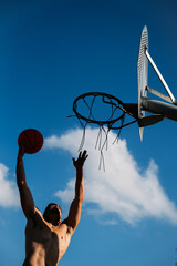 Young caucasian guy seen from below, muscular shirtless while jumping and hitting a basketball on a metal rack and clear blue sky on a sunny day. Dress the silhouette with open arms.