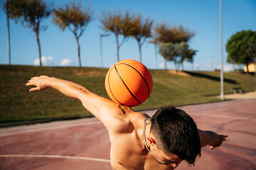 Muscular young shirtless Caucasian guy seen from close up while playing and juggling a basketball on a basketball court.