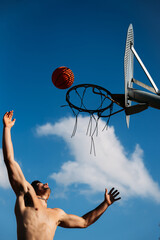Young caucasian guy seen from below, muscular shirtless while jumping and hitting a basketball on a metal rack and clear blue sky on a sunny day. Dress the silhouette with open arms.