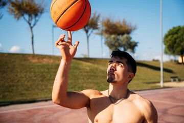 Muscular young shirtless Caucasian guy seen from close up while playing and spinning a basketball on a basketball court.