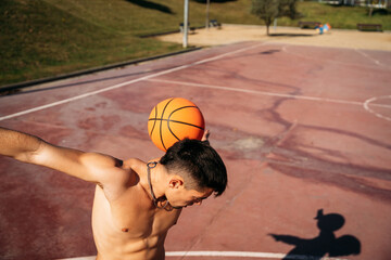 Muscular young shirtless Caucasian guy seen from close up while playing and juggling a basketball on a basketball court.