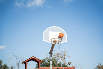 Basketball basket seen from below and one side while an orange ball is falling from above. Contrasted with a blue sky on a sunny, clear day.