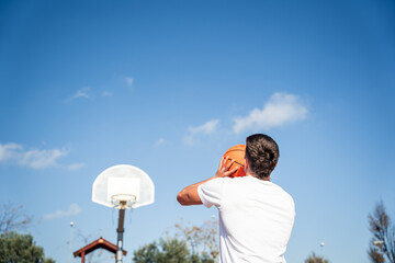 Young Caucasian boy with his back turned, wearing a white t-shirt throwing a basketball at a metal basket and the clear blue sky on a sunny day.