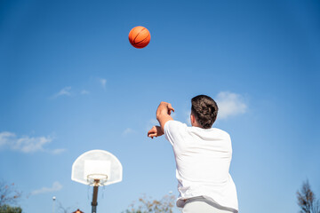 Young Caucasian boy with his back turned, wearing a white t-shirt throwing a basketball at a metal basket and the clear blue sky on a sunny day.
