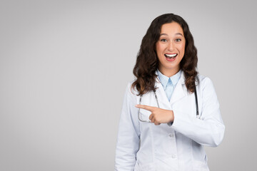 Shocked young lady doctor pointing finger aside at free space, standing on gray background, studio shot, banner