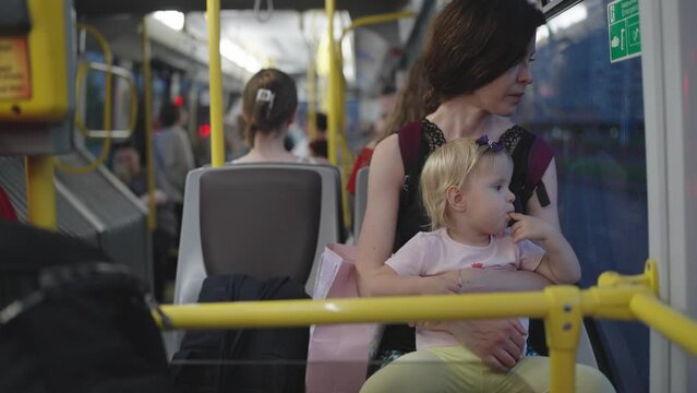 Mother and her little daughter are traveling on public transport in evening, looking out window. Calm pretty mother and daughter sitting by window in bus. A mother kisses her child with great love.