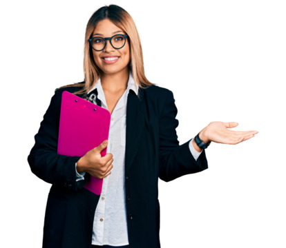 Young hispanic business woman holding clipboard with blank space smiling cheerful presenting and pointing with palm of hand looking at the camera.