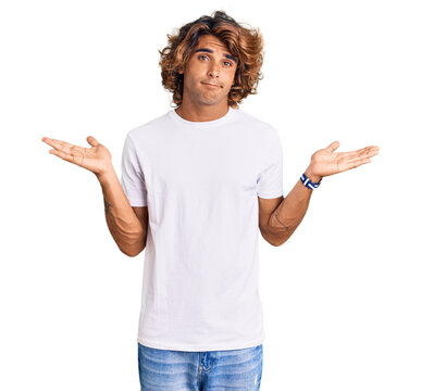 Young hispanic man wearing casual white tshirt clueless and confused expression with arms and hands raised. doubt concept.
